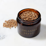 An Evermill brown glass jar labeled "Organic Coriander Seeds Refill" is filled with coriander seeds, highlighting their aromatic flavor profile. Several seeds are scattered on a white marble surface next to the jar. The lid is absent from the image, suggesting their journey from Bulgaria to your kitchen.
