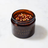 A brown glass jar labeled "Organic Red Pepper Flakes Refill" from Evermill, containing crushed red pepper flakes, sits on a light-colored marble surface. The open jar showcases its savory heat, making it a vibrant addition to any spice collection.