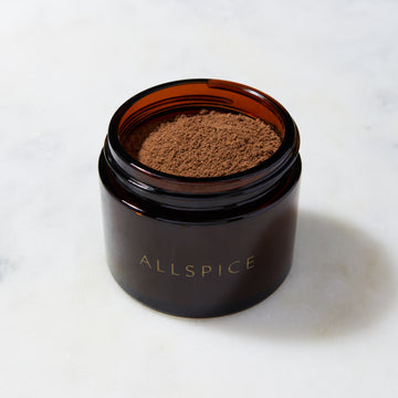 An attractively labeled glass jar marked "Organic Allspice Refill" from Evermill contains a finely ground allspice powder made from dried berries. The jar is open, revealing the aromatic spice inside, often sourced from the West Indies. It is placed on a pristine white marble surface.