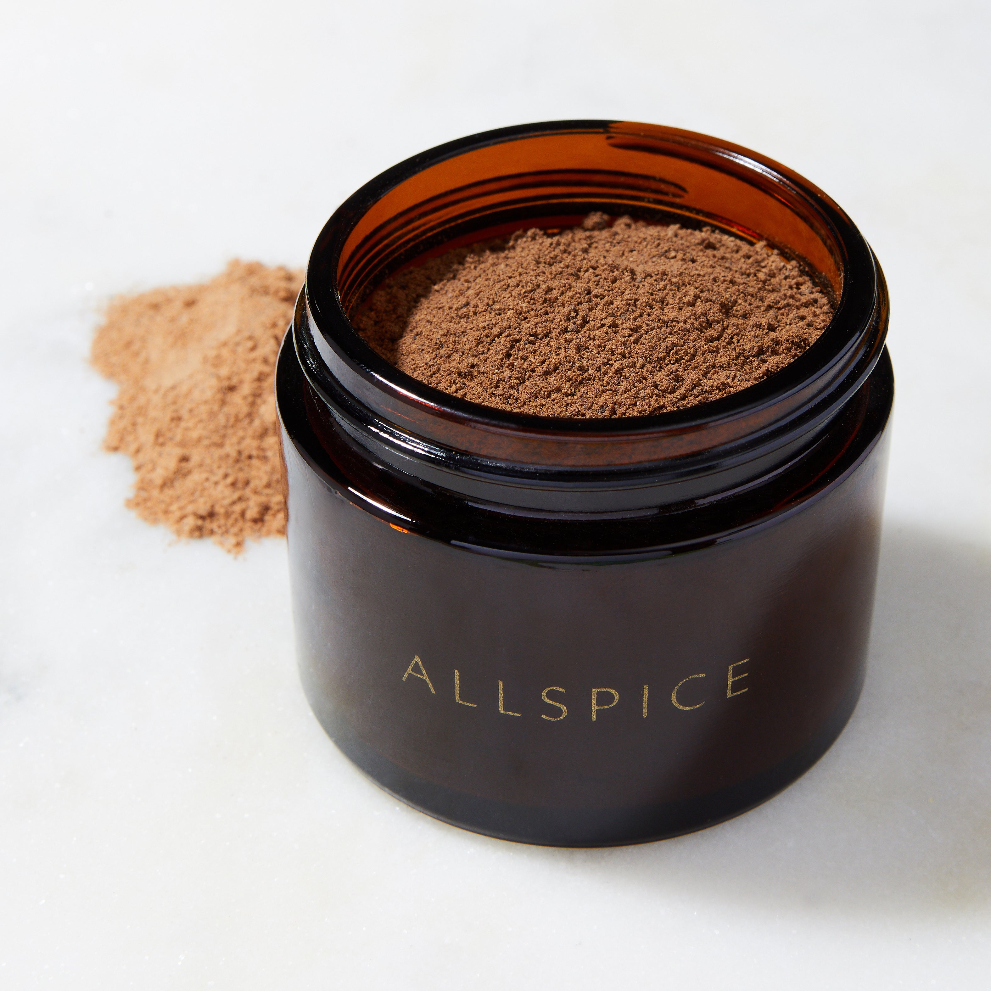 An open Evermill Organic Allspice Refill, in a brown glass jar, sits on a white surface. Some finely ground allspice powder made from the dried berries of the West Indies has spilled beside the jar.